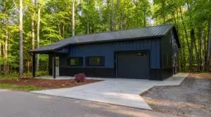 Navy blue steel garage with black roof and driveway in a forest setting.